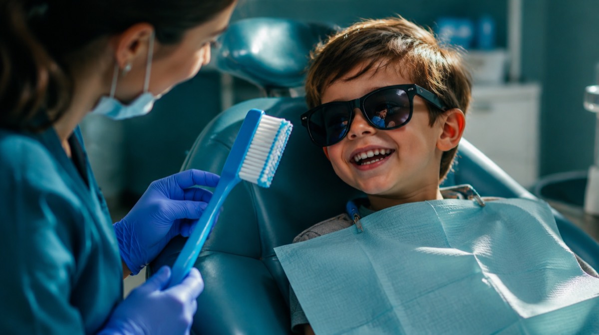 Young boy in sunglasses laughing in a dental chair while a pediatric dentist shows him a giant toothbrush